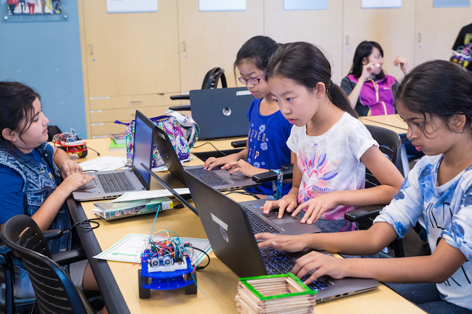 Four female middle school students in summer robotics academy are gathered around a desk. All four students are sitting in front of laptops and writing code for their robots.
